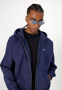 Navy blue zip-up hoodie with a drawstring hood, front pockets, and "OBEY" logo stitching, worn over a black shirt. Textured fabric.