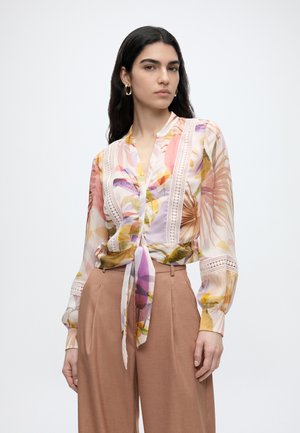 Frau mit langen dunklen Haaren, die goldene Creolen trägt, eine durchsichtige bunte Blumenbluse mit Spitzen-Details, die an der Taille gebunden ist, und hoch taillierte beige Hosen.