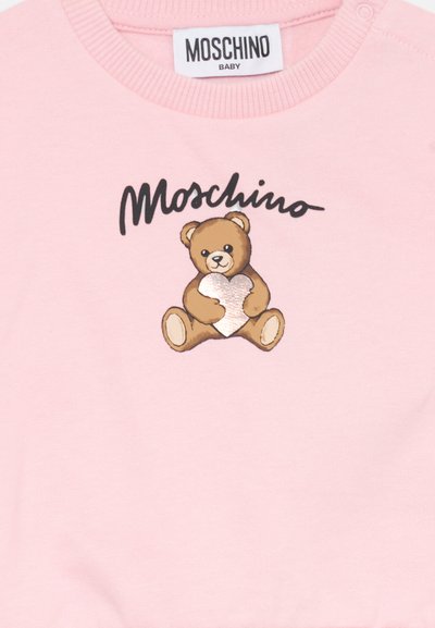Rožinis medvilnės džemperis su meškino grafika ir metaliniu širdies motyvu. Virš dizaino juodai užrašyta "Moschino Baby".