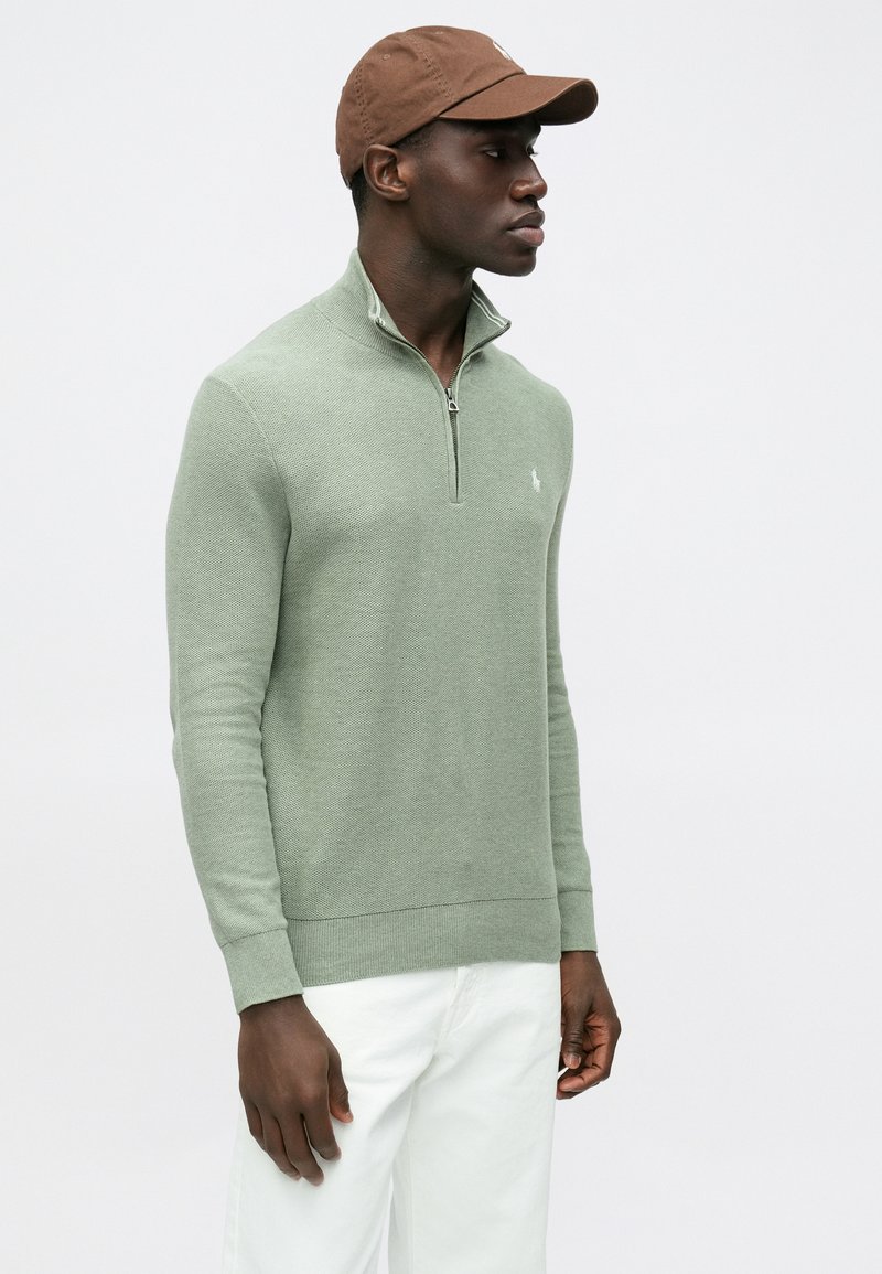 Maglione verde lavorato a maglia con collo a mezza zip, polsini a coste e piccolo logo sul petto. Abbinato a pantaloni bianchi e cappellino marrone.