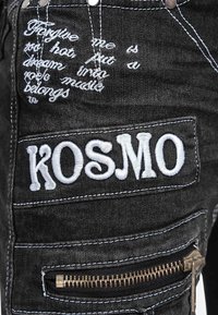 Black denim with white embroidered text reading "KOSMO" and cursive script, stitched seams, and a brass zipper detail.