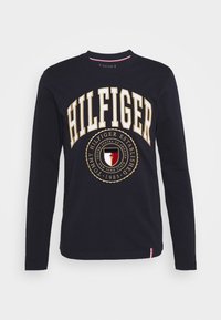 Laivaston pitkähiha t-paita, jossa on rohkea "HILFIGER"-teksti ja pyöreä logo punaisilla ja kultaisilla yksityiskohdilla edessä. Puuvillakangasta, pyöreä kaula-aukko.