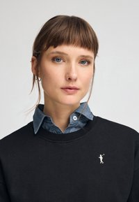 Svart sweatshirt med ribbad krage, lagd över en ljusblå denimskjorta. Litet vitt logotyp på bröstet. Slät tygtextur.