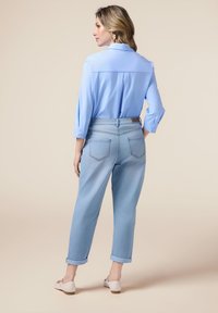 Camisa de botones azul claro, de corte relajado, combinada con jeans azul claro arremangados que cuentan con bolsillos traseros y un detalle de costura. Fondo neutro.