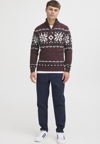 Maglione a maglia color bordeaux con motivo di fiocchi di neve bianchi e azzurri, collo a mezza zip, polsini a coste, abbinato a pantaloni blu navy e sneakers bianche.