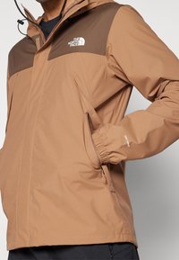 Veste imperméable marron avec un panneau supérieur marron contrastant, poignets élastiques, poches latérales et capuche. Logo visible sur la poitrine.