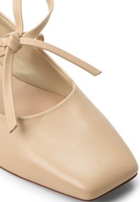 Beige Leder Schuh mit quadratischer Zehenpartie, glatter Oberfläche und einem dünnen Knöchelriemen, der zu einer Schleife gebunden ist. Mit einem niedrigen Blockabsatz.
