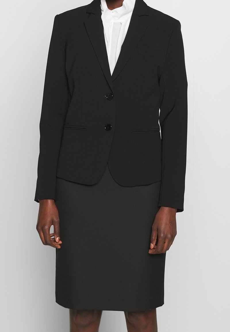Personne portant un blazer noir cintré sur un chemisier blanc et une jupe noire assortie jusqu'au genou, debout devant un fond uni.
