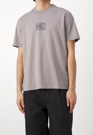 Homme portant un t-shirt gris clair à manches courtes avec un petit texte noir en miroir "ALLSAINTS" sur la poitrine et un pantalon noir, debout sur fond blanc.