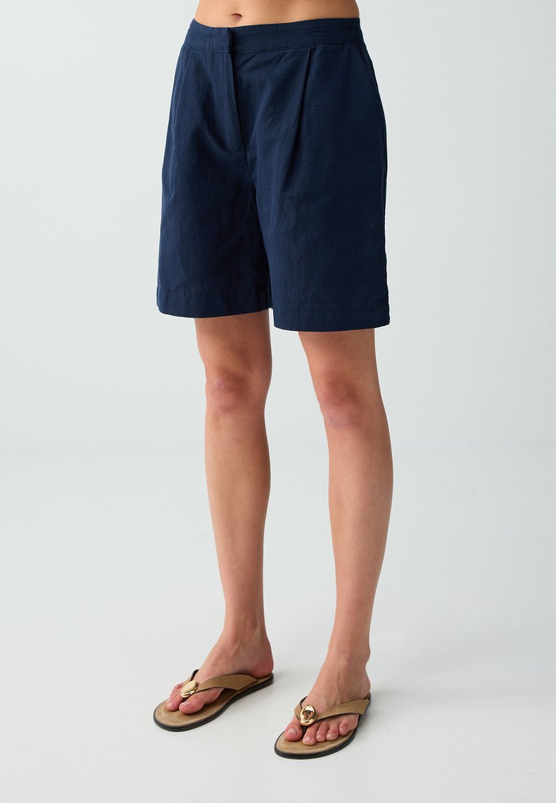 Personne portant un short bleu marine au genou et des tongs beige avec une décoration ronde dorée, debout devant un fond clair uni.