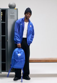 Kék Nike hátizsák dombornyomott logóval, cipzáras záródással és állítható pántokkal, egy kék dzsekis és fekete nadrágos személy által tartva.