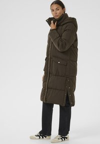 Polstret mørk olivengrøn frakke med hætte, sidelukknapper, store forlommer og knælængde. Båret over mørke jeans og sneakers.