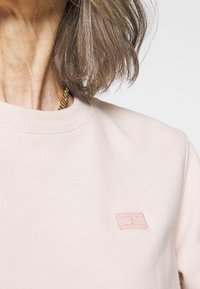 Felpa rosa chiaro con colletto rotondo, caratterizzata da un logo rettangolare ricamato di un rosa leggermente più scuro. Tessuto morbido.