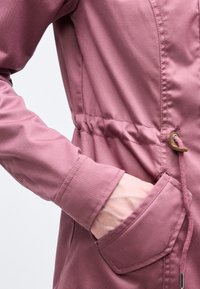 Χέρι ατόμου μέσα στην μπροστινή τσέπη ενός μωβ μακρυμάνικου μπουφάν με κορδόνι στη μέση και εμφανείς ραφές.