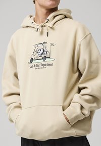 Beige hoodie featuring a front graphic of a cartoon character in a golf cart, with drawstrings and a large front pocket. Soft texture.