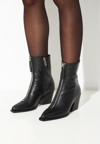Zwarte leren enkelboots met spitse neuzen en chunky hakken, voorzien van zilveren ritsen aan de buitenzijden en een gladde textuur.