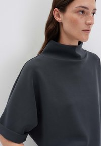 Donna con leggere lentiggini che indossa un top scuro a collo alto e maniche corte, rivolta a destra su uno sfondo chiaro e semplice.