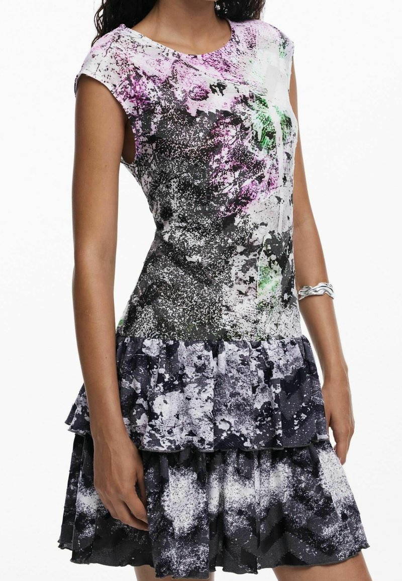Femme portant une robe sans manches avec un motif abstrait noir, blanc, violet et vert et une jupe à volants superposés, bracelet argenté au bras.