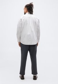 Άνδρας με πλεξούδες στέκεται με την πλάτη προς τα εμάς, φοράει ένα ανοιχτό πουκάμισο με ρίγες, σκούρο παντελόνι, λευκές κάλτσες και καφέ παπούτσια σε λευκό φόντο.