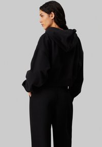 Felpa nera a maniche lunghe con cappuccio, caratterizzata da maniche ampie e una texture morbida. Abbinata a pantaloni neri coordinati. Vista posteriore.