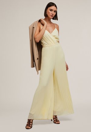 Une combinaison plissée jaune avec un décolleté en V et des bretelles fines, dotée d'une taille élastique. Assortie à un blazer beige et des talons marron à lanières.