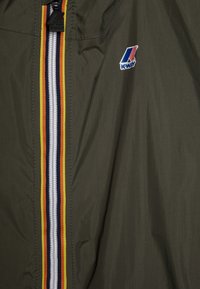 Veste légère verte avec le logo K-Way, dotée d'une fermeture éclair centrale avec des rayures jaunes, blanches et bleu marine. Tissu lisse et résistant à l'eau.