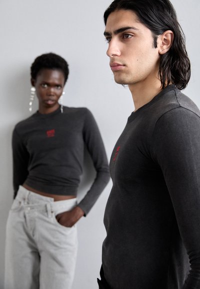 Graue Langarmshirts mit roter Stickerei, kombiniert mit hellgrauen, hochtaillierten Jeans. Weicher Stoff, figurbetontes Design, dezente Muster.