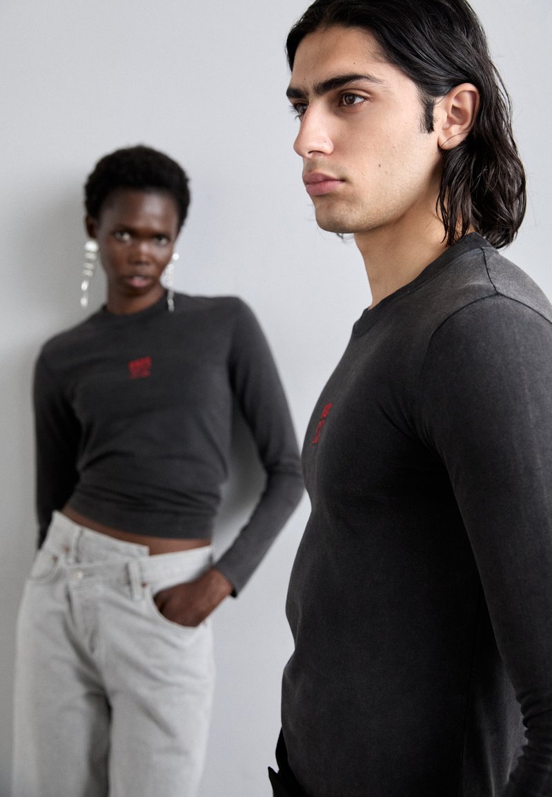 Graue Langarmshirts mit roter Stickerei, kombiniert mit hellgrauen, hochtaillierten Jeans. Weicher Stoff, figurbetontes Design, dezente Muster.