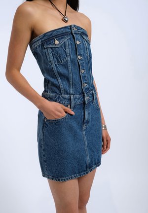 Abito di denim blu senza spalline con bottoni frontali e tasche, indossato da una persona con una collana con ciondolo a forma di cuore e una mano in tasca.