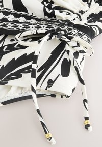 Tissu à motifs noir et blanc avec détails froncés et extrémités de cordon de serrage nouées ornées de perles dorées sur fond clair.