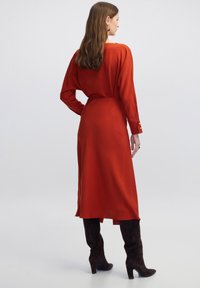Vestido largo rojo con un brillo sutil, mangas largas, cintura entallada y abertura lateral. Combinado con botas de ante oscuras hasta la rodilla.