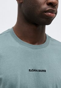 Κοντινό πλάνο ενός ατόμου που φοράει μπλουζάκι με στρογγυλή λαιμόκοψη σε τιρκουάζ χρώμα με μαύρο κεντημένο κείμενο στο κέντρο του στήθους.