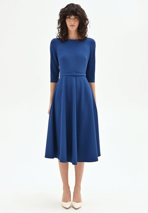 Femme en robe bleu royal mi-longue avec manches trois-quarts et ceinture assortie, debout devant un fond blanc uni.