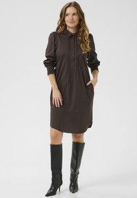 Robe marron à col et manches longues avec patte de boutonnage, dotée de poches latérales et de poignets froncés ; associée à des bottes noires élégantes jusqu'au genou.