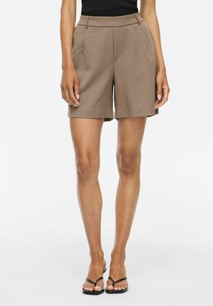 Femme portant un short taupe taille haute ajusté avec des plis, un haut noir et des sandales noires type tongs, debout sur un fond blanc.