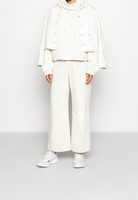 Biała skórzana kurtka na krótkim rękawie noszona na puszystym białym hoodie, zestawiona z szerokimi białymi spodniami. Strój charakteryzuje się miękką teksturą i casualowym stylem, podkreślonym białymi sneakersami.