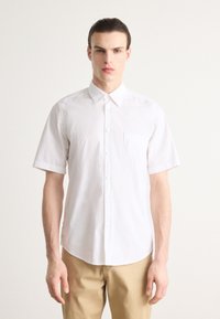 Camisa blanca de manga corta con cuello puntiagudo, bolsillo en el pecho y una textura suave, combinada con pantalones beige claros.