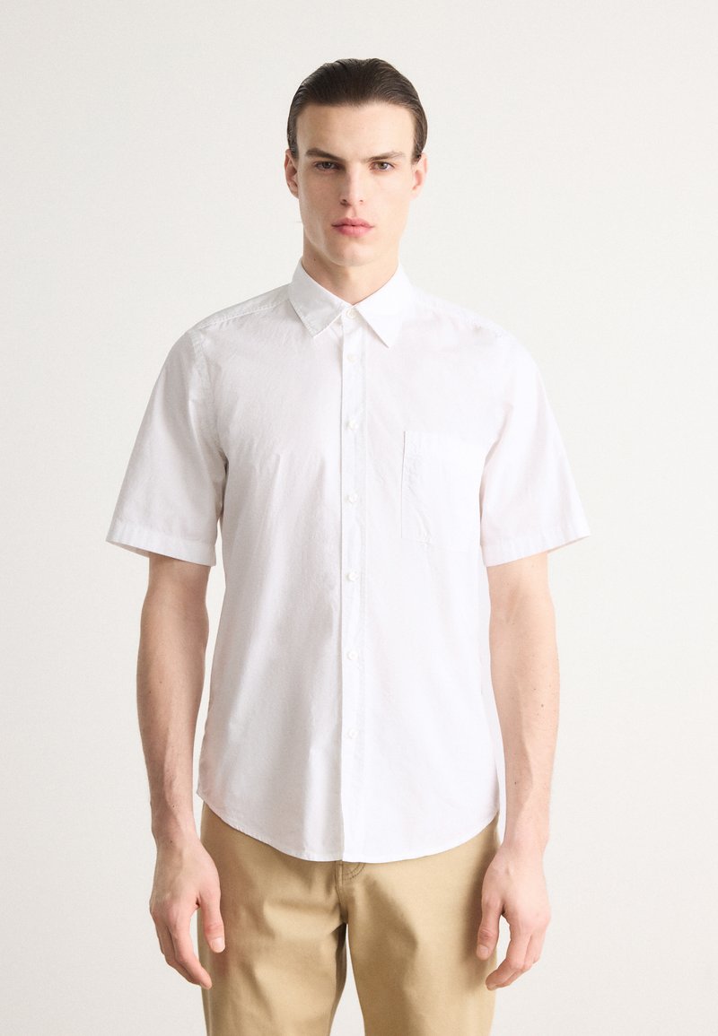 Camisa blanca de manga corta con cuello puntiagudo, bolsillo en el pecho y una textura suave, combinada con pantalones beige claros.