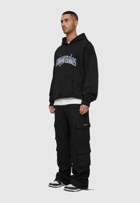 Sort oversized hoodie med grafisk tekst, parret med sorte cargo bukser med flere lommer og en afslappet pasform. Sneakers med hvide og sorte accenter.
