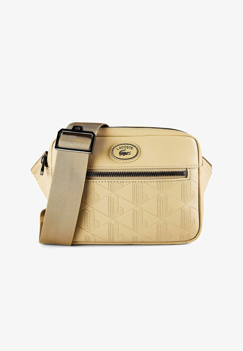 Sac bandoulière beige Lacoste avec motif géométrique en relief, poche avant zippée, sangle large réglable et patch logo ovale.