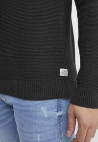 Zwarte gebreide trui met textuurstof en geribde manchetten, voorzien van een klein merklabel aan de onderzijde, gecombineerd met lichtblauwe jeans.