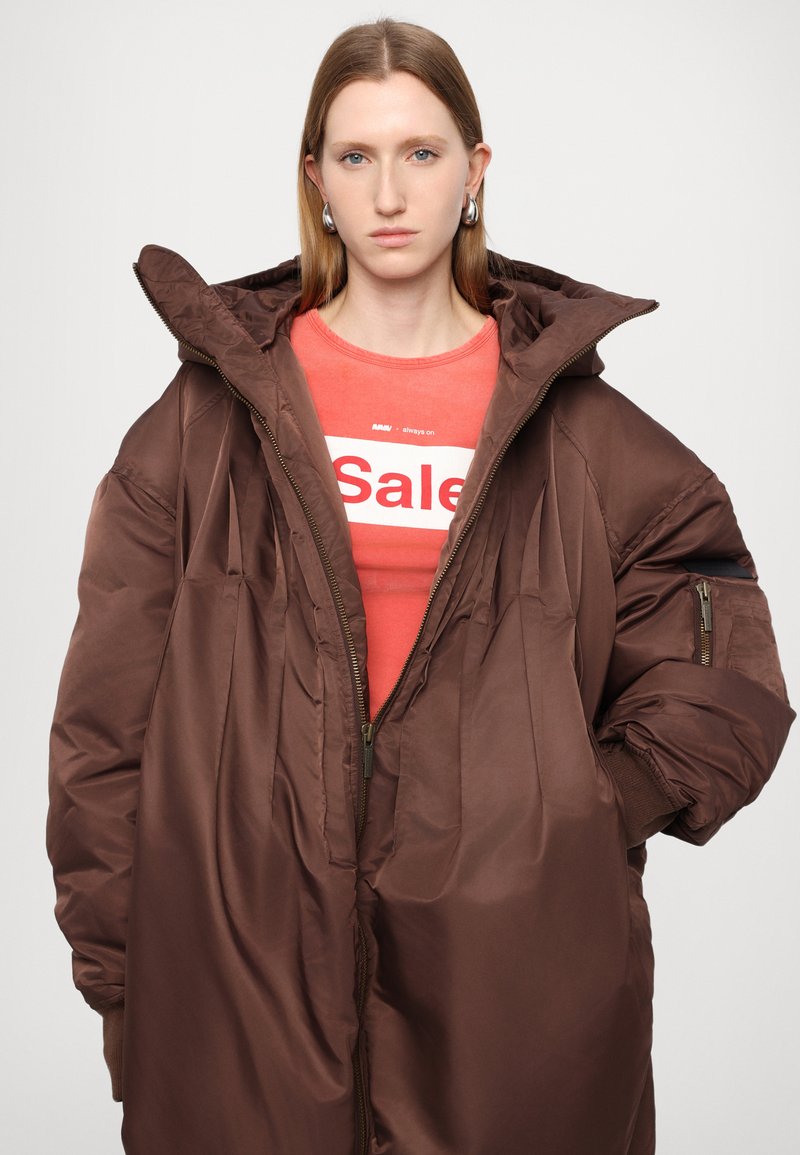 Brauner Puffermantel mit Kapuze, mit Reißverschluss, Seitentaschen und gerippten Bündchen. Darunter ein rotes T-Shirt mit "Sale" in Weiß bedruckt.