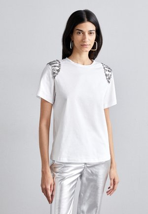 T-shirt bianco in cotone a maniche corte, con dettagli in argento sulle spalle. Indossato con pantaloni argentati lucidi.