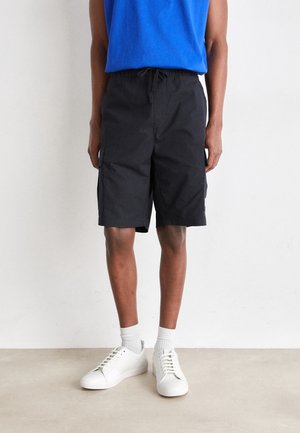 Person iført sorte cargoshorts, hvide sokker, hvide sneakers og en blå ærmeløs skjorte, stående mod en ensfarvet hvid væg.