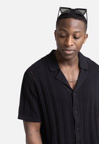 Camisa negra de mangas cortas con un patrón texturizado y cierre de botones en el frente, que cuenta con un collar y sutiles detalles en corte.