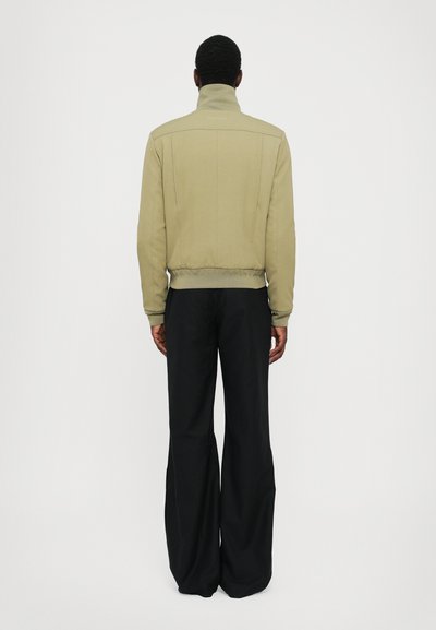 Beige bomber jacket with high collar and ribbed hem, featuring visible seams and minimalistic design, paired with loose black pants.