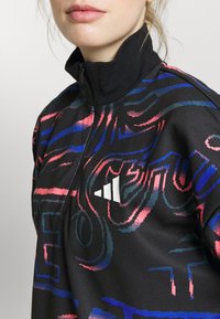 Femme portant une veste noire à fermeture quart zip avec des motifs abstraits roses et bleus et un logo blanc Adidas sur la poitrine.