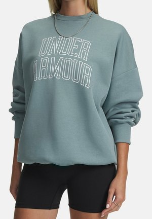 Vrouw draagt een losse lichtblauwe Under Armour sweatshirt, zwarte biker shorts, zilveren ketting en ringen, staat met ontspannen armen.