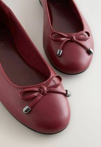 Ballet flats nahkaa syvänpunaisena, pyöreällä varvalla, jossa on nauha detalji ja metalliset korostukset nauhojen päässä. Sujuva pinta.