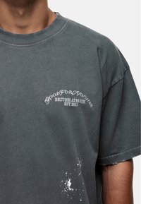 Donkergrijs oversized T-shirt van katoen, met een ronde halslijn, borduursel aan de voorkant en witte verfspetterdetails aan de onderzijde.